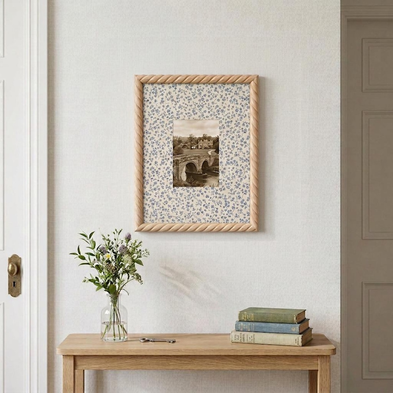 May include: A rectangular picture frame with a light wood finish and a twisted rope texture border. The frame contains a mat with a blue and white floral pattern surrounding a central black and white photograph of a stone bridge and building.