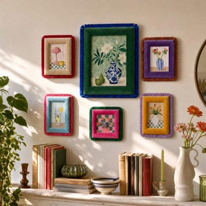 Op de afbeelding: Een galerijwand met zes rechthoekige fotolijsten met zachte fluwelen randen in rood, groen, blauw, paars en roze. De lijsten bevatten diverse botanische illustraties en stillevens, opgehangen boven een boekenplank met verschillende boeken.