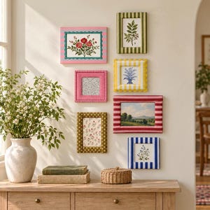 May include: A collection of seven rectangular picture frames with varied patterns and colors, including stripes, dots, and solid textures, arranged on a wall. The frames contain botanical prints, landscape art, and patterned paper inserts.
