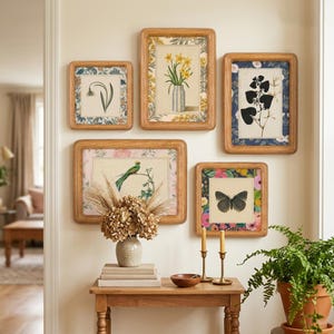 May include: A collection of five framed botanical prints with wooden frames. The prints feature various floral and fauna illustrations, including daffodils, a bird, and a butterfly. The frames are mounted on a cream-colored wall above a wooden table.