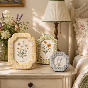 May include: Three scalloped-edge picture frames featuring botanical illustrations. The largest frame has a yellow floral pattern with a chamomile print. The medium frame has a green floral pattern with a calendula print. The smallest has a blue striped pattern with a lavender print.