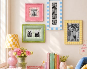 May include: A collection of four rectangular picture frames in pink, light blue, sage green, and yellow, featuring beaded or checkered patterns. The frames display black and white photographs of people, arranged on a wall above a table with decor.