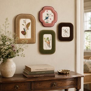 Op de afbeelding: Vier wandframes met fluwelen randen in beige, roze, groen en donkerbruin, elk met een botanische vogelillustratie. Daaronder staat op een houten sidetable een keramieken vaas met wilde bloemen en drie op elkaar gestapelde boeken.