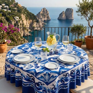 May include: A round table covered with a white tablecloth featuring a blue floral and scroll pattern. The table is set with white plates, clear wine glasses, a glass pitcher of lemonade with lemon slices, and small blue and white ceramic shakers.