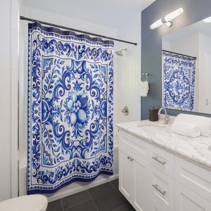 May include: A shower curtain featuring a blue and white pattern with intricate scrollwork, floral motifs, and central fruit illustrations. The curtain hangs from a black rod and is displayed in a bathroom with a white vanity and a mirror.