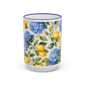 Op de afbeelding: Witte keramische mok met een blauwe rand, versierd met een bloemenpatroon van blauwe hortensia's en gele citroenen op een crèmekleurige achtergrond. Het ontwerp bevat groene bladeren en kleine witte bloemen.