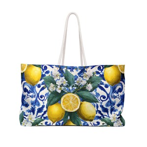 May include: A large tote bag featuring a vibrant pattern of bright yellow lemons, green leaves, and white blossoms set against a blue and white decorative scrollwork background. The bag includes two white rope handles attached to the top edge.