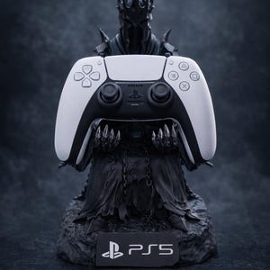 Op de afbeelding: Een zwart beeldje van een donker gepantserde figuur met een stekelige kroon die een witte en zwarte draadloze gamecontroller vasthoudt. De basis van het beeldje heeft een rechthoekig plaatje met het witte PlayStation-logo en de tekst PS5.