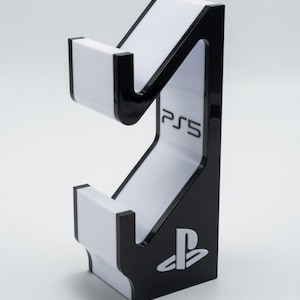 Pode incluir: Um suporte de console de jogos PS5 em preto e branco. O suporte apresenta o logotipo PS5 e o símbolo PlayStation. O suporte foi projetado para conter um console de jogos e acessórios. O suporte é feito de um material brilhante.