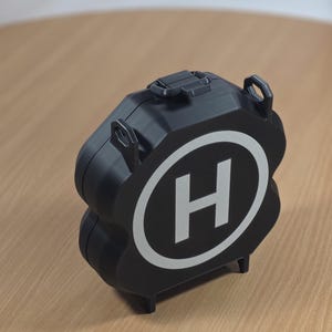 Pode incluir: Recipiente preto multicamadas com um "H" branco dentro de um oval branco, assemelhando-se a um símbolo de heliporto. O recipiente tem uma alça e duas alças. Está sobre uma superfície de madeira castanha clara.