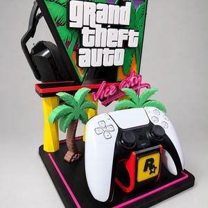 Op de afbeelding: Een zwarte, gele en groene gaming displaystandaard met het Grand Theft Auto Vice City-logo, twee palmbomen en een Rockstar Games-embleem. De standaard houdt een wit-zwarte gamecontroller en een paar zwarte hoofdtelefoons vast.