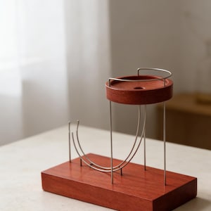 Pode incluir: Um suporte de madeira moderno e minimalista, com acabamento castanho-avermelhado. Possui uma base retangular e um topo circular, conectados por finos suportes metálicos curvos. O topo tem uma pequena reentrância circular.