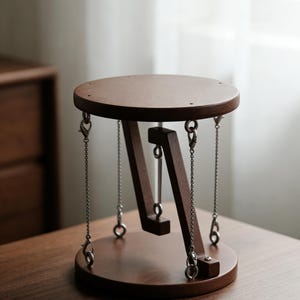 Op de afbeelding: Een bruine houten bijzettafel met een cirkelvormig blad en voet, verbonden door schuine steunen. De bovenkant wordt opgehangen door metalen kettingen. De tafel staat op een houten oppervlak.