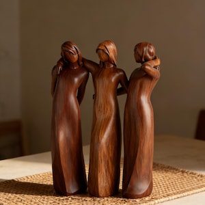 Könnte beinhalten: Eine Holzskulptur, die drei Figuren in langen Kleidern darstellt, jede mit einem Arm um die andere. Die Figuren sind aus dunklem, rotbraunem Holz geschnitzt und stehen auf einer gewebten Matte. Die Skulptur ist ein dekorativer Gegenstand.