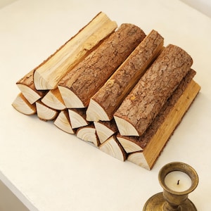 May include: A stack of firewood logs with rough, brown bark and light-colored wood ends. The logs are arranged in a triangular shape. A small, antique brass candle holder with a lit white candle sits nearby.