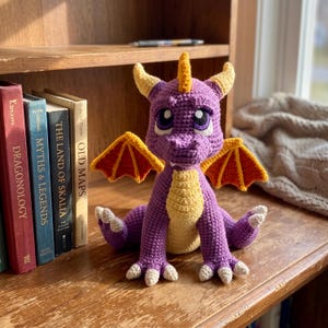 Pode incluir: Um peluche de dragão em croché roxo com peito amarelo, chifres amarelos e asas cor de laranja com rebordo amarelo. O dragão tem olhos roxos grandes com reflexos brancos e pequenas garras brancas nos pés, sentado numa posição vertical.