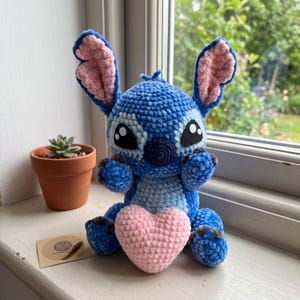 Pode incluir: Um peluche de croché azul com orelhas grandes com detalhes interiores cor-de-rosa, olhos pretos e brancos e um nariz azul escuro. O peluche está sentado e segura um coração de croché cor-de-rosa claro contra o peito, feito de fio macio.