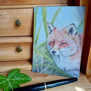 Könnte beinhalten: Ein detailliertes Aquarellgemälde eines Rotfuchses mit hellblauem Hintergrund und grünem Gras. Der Fuchs hat eine weiße Brust und einen fokussierten Blick. Das Kunstwerk wird auf einer Holzoberfläche präsentiert.