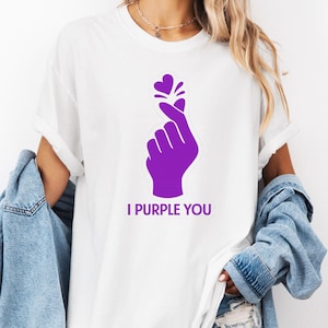 May include: White t-shirt with a purple graphic of a hand making a heart shape, with the text "I PURPLE YOU" below. The design features a heart symbol above the fingers. The shirt is a casual style.