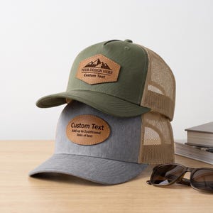 Op de afbeelding: Twee truckerpetten verticaal op elkaar gestapeld. De bovenste pet is olijfgroen met een beige mesh achterkant en een zeshoekige leren patch met een berggrafiek en de tekst YOUR DESIGN HERE Custom Text. De onderste pet is grijs gemêleerd met een beige mesh achterkant en een ovale leren patch met de tekst Custom Text Add up to 2additional lines of text.