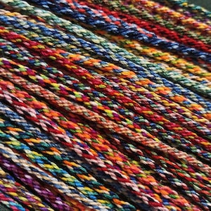 May include: A collection of multiple braided cords featuring a vibrant, multi-colored pattern. Each cord is composed of tightly twisted threads in various shades of red, blue, yellow, green, orange, and purple, creating a textured, rope-like appearance.