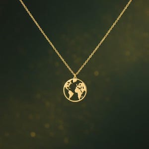 Puede incluir: Un collar dorado con un colgante con forma de mapa del mundo. El colgante es un contorno circular con los continentes recortados. La cadena es delicada y también dorada, sobre un fondo verde oscuro.