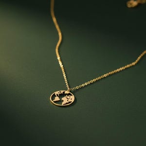 Puede incluir: Un collar dorado con un colgante en forma de mapa del mundo. El colgante ovalado presenta continentes recortados. La fina cadena cuelga sobre un fondo verde oscuro. Una joya elegante.
