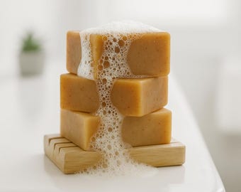 May include: A stack of four rectangular, light brown soaps on a wooden soap dish. The soaps are covered in white, foamy bubbles. The soap dish has a slotted design.