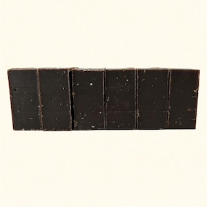 May include: A close-up shot of seven dark chocolate bars arranged side by side. The bars are rectangular, with a slightly rough texture and visible flecks. The chocolate is a deep, rich brown color.