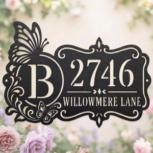 May include: A black metal house sign featuring a large letter B, the number 2746, and the text WILLOWMERE LANE. The sign is decorated with two butterfly silhouettes and ornate scrollwork, with a light-coloured background showing through the cutouts.