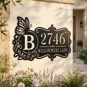 Pode incluir: Placa de número de casa em metal preto com design de borboleta. A placa exibe o número 2746 e o nome da rua Willowmere Lane. A letra "B" é apresentada em um círculo com uma borboleta. A placa é montada em uma parede bege.