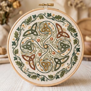 Puede incluir: Un bastidor de bordado circular de madera que contiene una tela de color blanco roto con un diseño bordado a mano. El patrón presenta un motivo central de nudo celta en tonos verde, dorado y óxido, rodeado por un borde de hojas de hiedra.