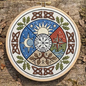 Puede incluir: Arte mural bordado con un diseño circular que representa elementos celestiales y naturales. La obra de arte incluye un sol, una luna, estrellas, una brújula, un árbol y olas, todo dentro de un bastidor de bordado de madera. La paleta de colores incluye azul, rojo, verde y marrón.