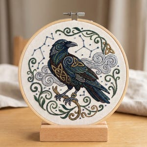 May include: A circular embroidery hoop containing a fabric panel with a stitched raven design. The raven features blue, black, and gold Celtic knotwork patterns, surrounded by embroidered grey clouds, green vines, and blue star constellations.