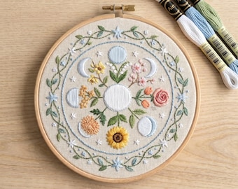May include: A round embroidery hoop featuring a floral and moon phase design. The embroidery includes a sunflower, roses, and other flowers in shades of yellow, pink, and blue. Several spools of embroidery floss in white, blue, and green are placed next to the hoop.
