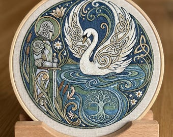 May include: A circular embroidery hoop containing a detailed needlework design of a white swan on blue water, a knight in silver armor holding a sword, and a tree of life motif, all framed by intricate Celtic knotwork and floral patterns.