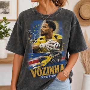 Könnte beinhalten: Ein anthrazitfarbenes T-Shirt in Acid-Wash-Optik mit einem Grafikdruck eines Fußballtorwarts im gelben Trikot. Das Design zeigt den Namen VOZINHA, den Text CABO VERDE, eine Inselkarte, Sterne und einen Fußball. Das Shirt hat einen Rundhalsausschnitt.