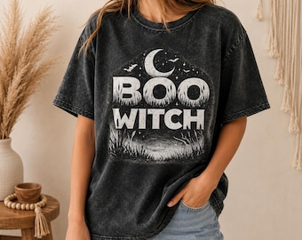 May include: A charcoal grey mineral-washed t-shirt featuring a white graphic print of a crescent moon, stars, and bats above the text "BOO WITCH" in a bold, distressed font, with a silhouette of tall grass at the base of the design.