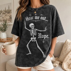 May include: A charcoal grey acid-wash t-shirt featuring a white graphic of a skeleton in a dancing pose. The text "Hear me out..." is printed above the skeleton, and the word "Nope." is printed below it in a decorative white serif font.
