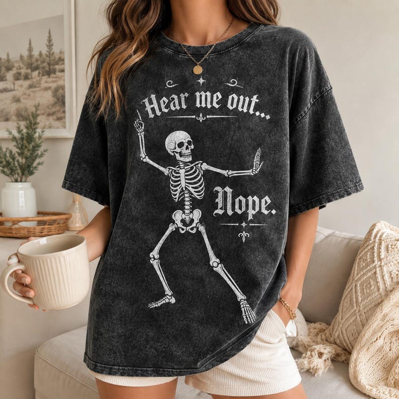 May include: A charcoal grey acid-wash t-shirt featuring a white graphic of a skeleton in a dancing pose. The text "Hear me out..." is printed above the skeleton, and the word "Nope." is printed below it in a decorative white serif font.