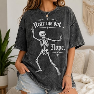 May include: A charcoal grey acid-wash t-shirt featuring a white graphic of a skeleton in a dancing pose. The text "Hear me out..." is printed above the skeleton, and the word "Nope." is printed below it in a decorative, vintage-style font.