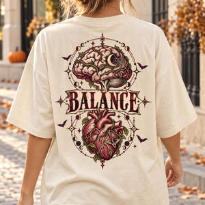 Könnte beinhalten: Ein beiges T-Shirt mit einem Rückendruck, der oben ein menschliches Gehirn und unten ein menschliches Herz zeigt, beide mit Ranken durchzogen. Das Wort BALANCE ist mittig zwischen den Organen gedruckt und von Mondphasen umgeben.