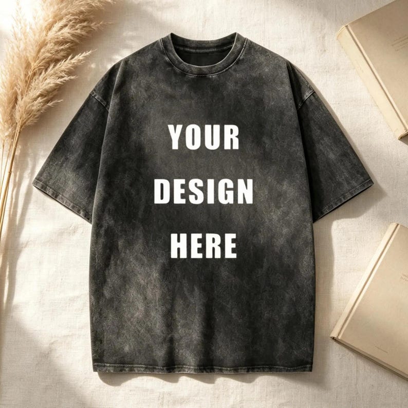 May include: A charcoal grey short-sleeve t-shirt with a mineral wash finish. The center of the chest features the white text "YOUR DESIGN HERE" in a bold, sans-serif font. The garment has a crew neckline and a relaxed, boxy fit silhouette.