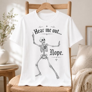 May include: A white short-sleeved t-shirt featuring a black graphic of a skeleton in a dynamic pose. The text "Hear me out..." is printed above the skeleton, and the word "Nope." is printed to the right of the skeleton in a decorative font.