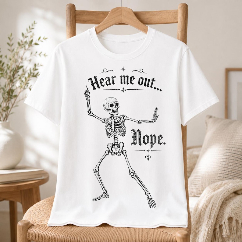 May include: A white short-sleeved t-shirt featuring a black graphic of a skeleton in a dynamic pose. The text "Hear me out..." is printed above the skeleton, and the word "Nope." is printed to the right of the skeleton in a decorative font.