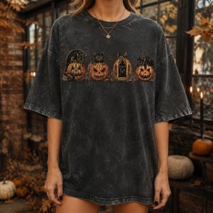 May include: A charcoal grey acid-wash t-shirt featuring a graphic print of four black cats perched on or near carved jack-o'-lantern pumpkins. The design includes intricate details of the cats and pumpkins arranged in a horizontal row across the chest.
