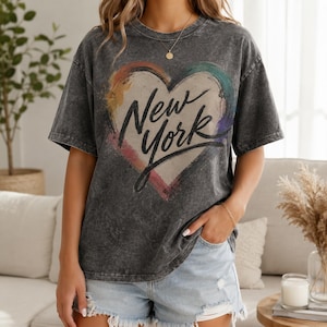 Op de afbeelding: Een antracietgrijs acid-wash T-shirt met een grote hartvormige afbeelding met een veelkleurige penseelstreekrand en de tekst New York in zwarte letters in het midden. Het T-shirt heeft korte mouwen en een ronde halslijn.