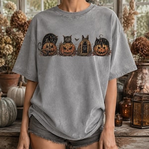 May include: A grey short-sleeve t-shirt featuring a graphic print of four black cats perched on or inside carved orange jack-o'-lanterns. The design includes a small moth between the cats and pumpkins, all set against a muted, washed-out grey fabric.