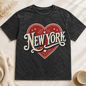 Peut inclure: Un t-shirt gris anthracite délavé à l'acide avec un grand cœur rouge imprimé sur la poitrine. À l'intérieur du cœur, le texte NEW YORK est inscrit dans une police vintage stylisée de couleur crème, entouré de lignes et de petites étoiles.