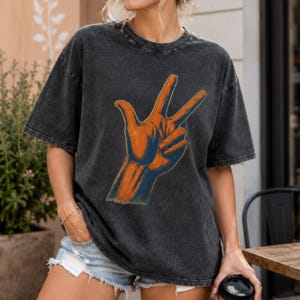 May include: A charcoal grey acid-wash t-shirt featuring a graphic print of a hand making a three-finger gesture. The hand illustration is rendered in shades of orange and blue with detailed cross-hatching lines throughout the design.
